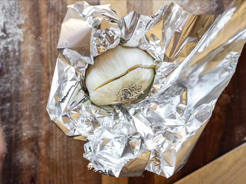 A bulb of garlic partially wrapped in crinkled aluminum foil, resting on a wooden surface.